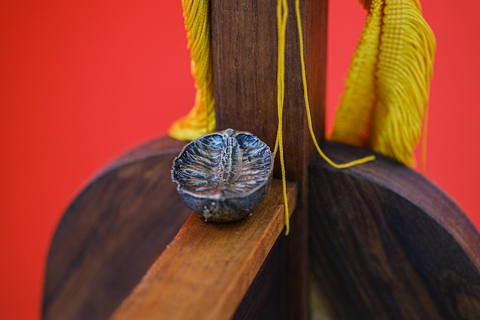 A silver looking metal casting of half a lime sits on a dark wooden structure. Behind drapes a yellow tassel against a bright red wall.
