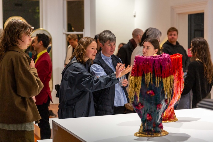 In a busy gallery four people gather around a white plinth laughing while holding wine glasses. Displayed on the plinth is a vessel of vibrant colours and playful textures.